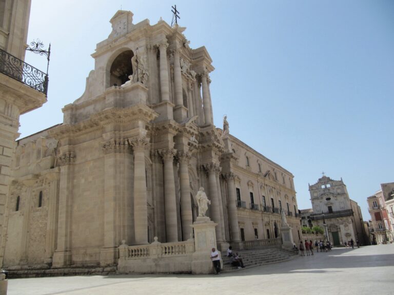 Facciata e fianco della Cattedrale di Siracusa su piazza Duomo, con Santa Lucia alla Badia sullo sfondo.